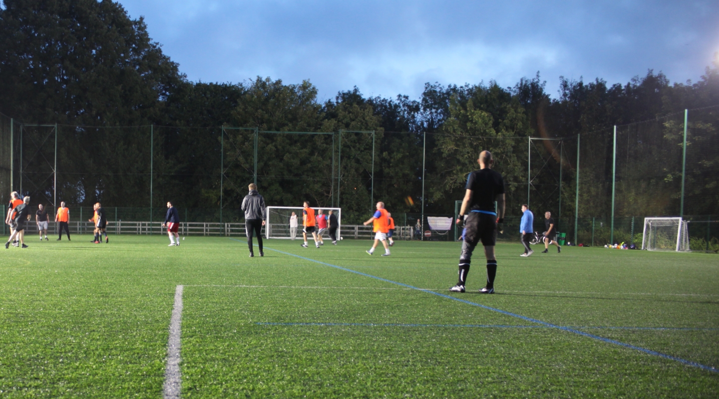 Coaches look on as players in orange bibs play football under floodlights at dusk.')