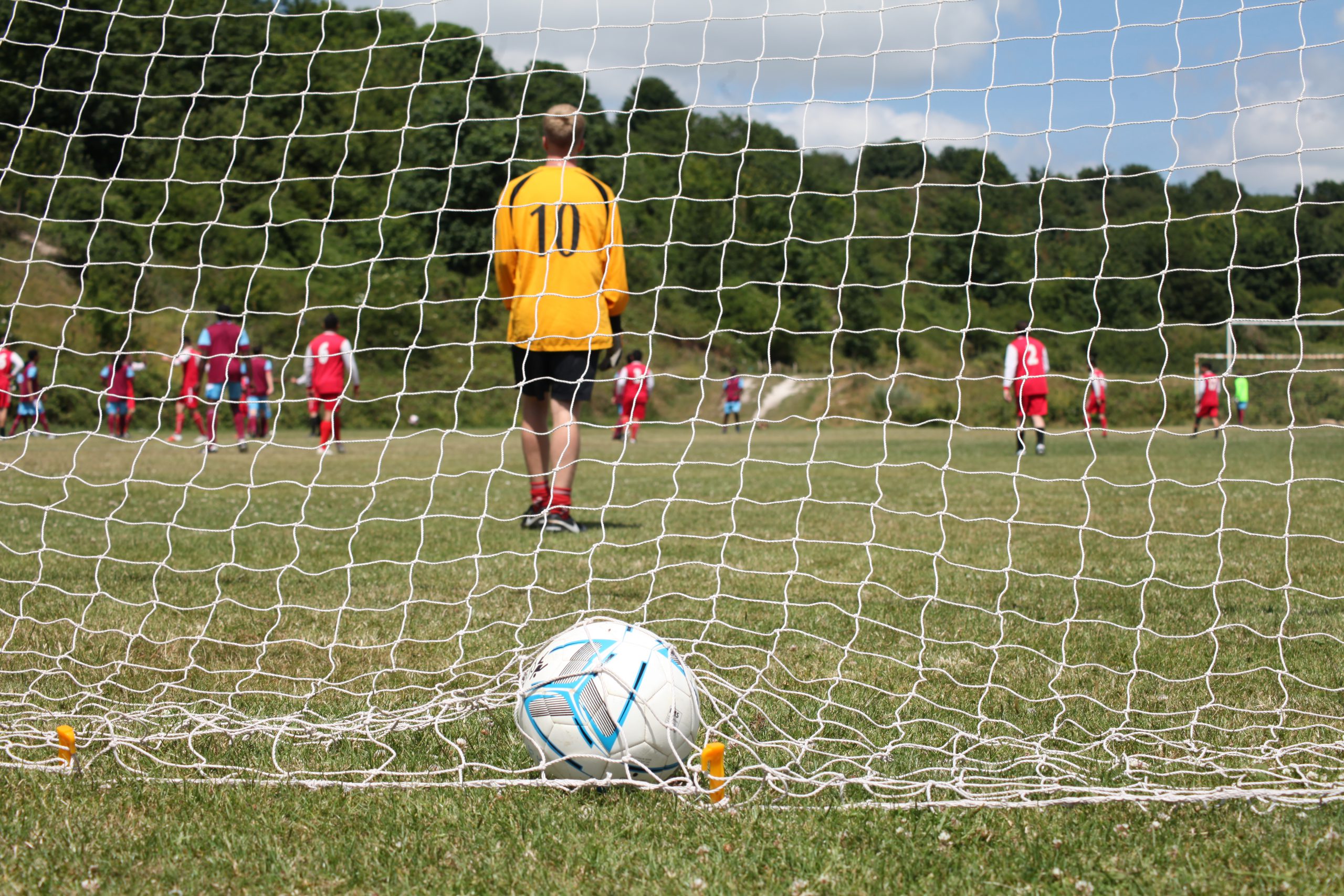Photo of a football in a goal net, taken from behind the goal with the back of a player in a yellow goalkeeper shirt looking away from us')
